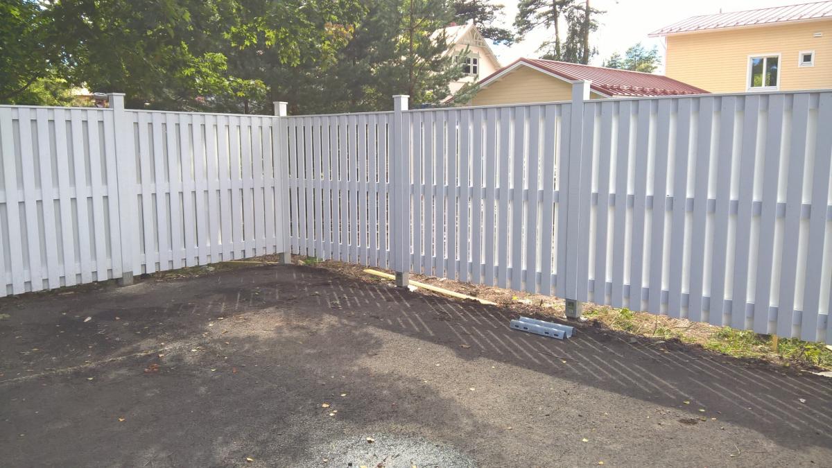 Valkoinen puuaita kulmassa aurinkoisena päivänä. Aidan takana näkyy vihreitä puita ja keltaisia taloja punaruskeilla katoilla. Maassa on tummaa asfalttia, jossa näkyy aidan pystysäleiden pitkiä varjoja. Aidan juurella on joitakin rakennustarvikkeita.