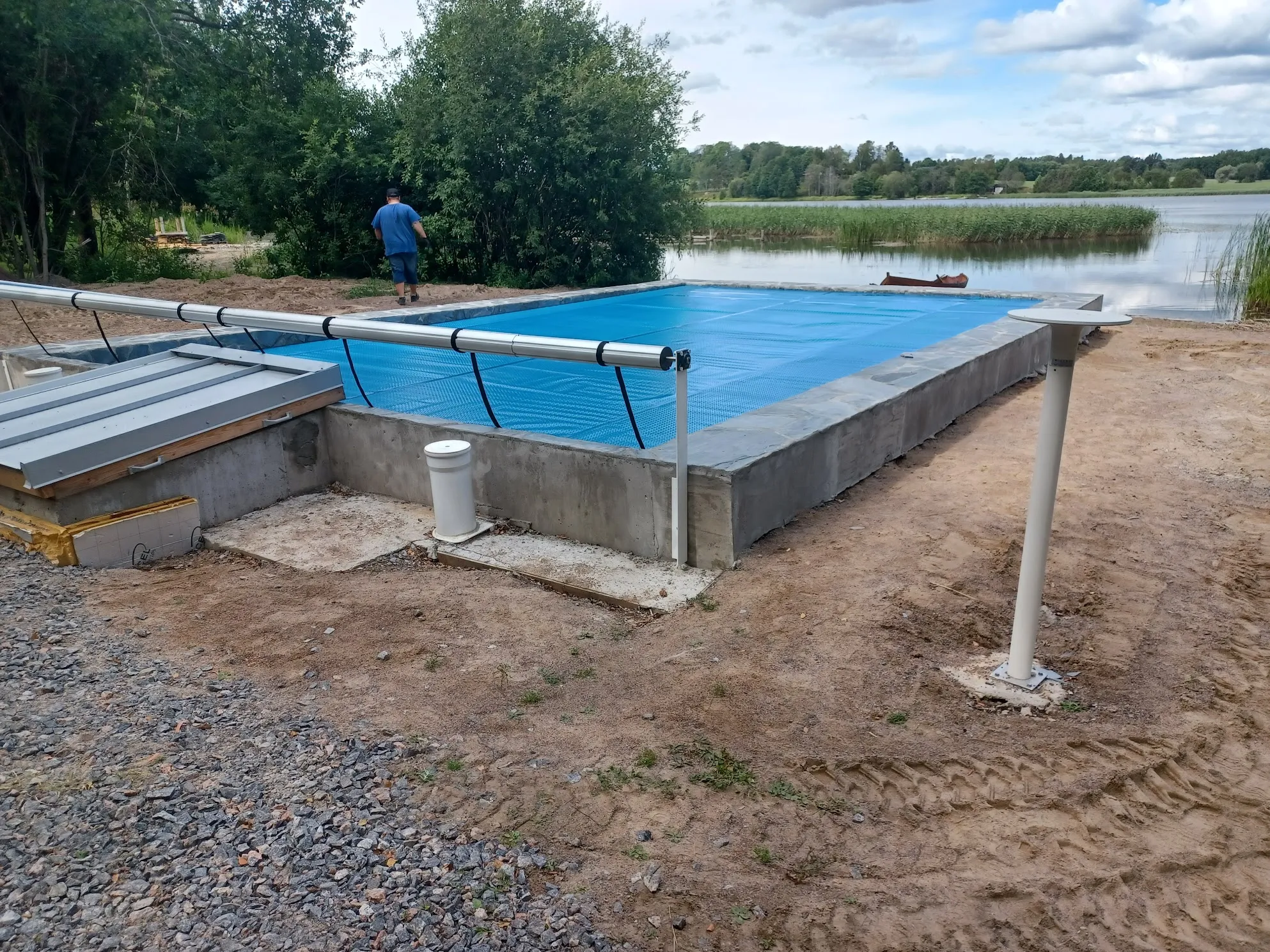 Keskeneräinen suorakaiteen muotoinen uima-allas, peitettynä kirkkaansinisellä kuplapeitteellä, seisoo hiekka- ja sorapohjaisella maalla. Altaan betonireunat ovat näkyvissä. Taustalla avautuu järvimaisema ruoikkoineen ja metsäisillä rannoilla pilvisen taivaan alla. Kuvassa mies kävelee poispäin altaasta kohti puita. Altaan vieressä on metallinen kansi ja valkoinen pylväs.