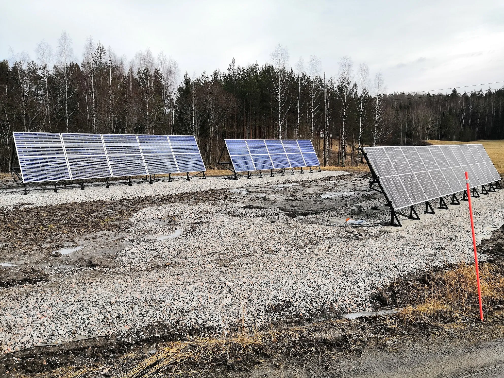 Kolme rivistöä aurinkopaneeleita seisoo soraisella ja mutaisella maalla. Kaksi vasemmanpuoleista rivistöä ovat sinisiä ja oikeanpuoleinen rivistö on harmaa. Taustalla on tiheä metsä, jossa on havupuita ja paljaita koivuja. Kaukana oikealla näkyy pieni punainen talo. Maassa on kuivaa ruohoa ja oikealla oranssi merkkitolppa. Taivas on pilvinen.