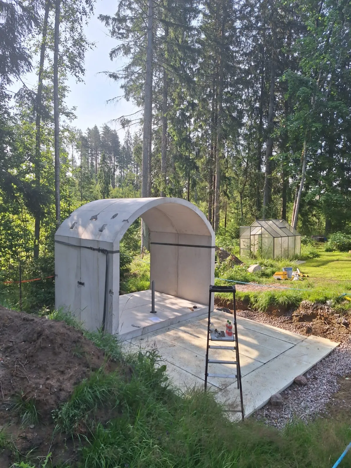 Vaaleanharmaa, kaarevakattoinen betonirakennelma seisoo metsän reunalla aurinkoisessa säässä. Sen edessä on kaivettua maata ja vihreää ruohoa. Oikealla puolella on mustat tikkaat ja taustalla näkyy tiheä puusto sekä pieni kasvihuone.