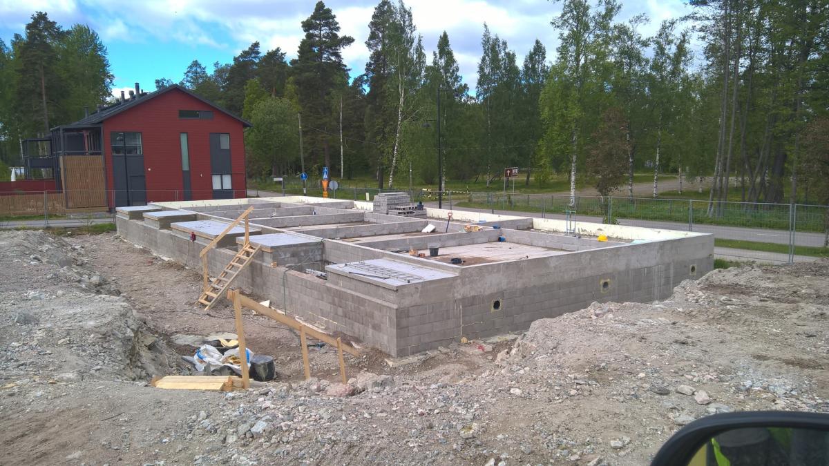 Etualalla rakennustyömaa, jossa on harmaa betoniharkoista rakennettu talon perustus. Perustuksessa on useita osastoja ja siihen nojaa puiset tikkaat. Maaperä on soraista ja multaa. Taustalla vasemmalla on valmis punainen talo tummanharmailla ikkunoilla ja katolla. Kauempana näkyy tie ja tiheä metsä. Taivas on sininen ja pilvinen.