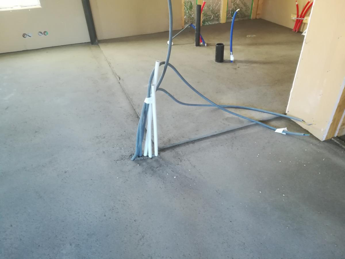 Kuva esittää rakenteilla olevan huoneen betonilattiaa. Lattian keskeltä nousee nippu harmaita ja valkoisia sähköputkia, ja harmaita kaapeleita kulkee lattiaa pitkin. Taustalla näkyy seinä, jossa on ikkuna ja sähkörasioita. Oikealla lattiasta nousee punaisia, sinisiä ja mustia LVI-putkia, ja seinää vasten nojaa rakennuslevy.