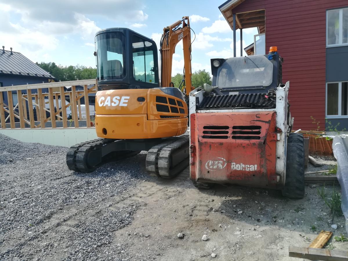 Kaksi rakennuskonetta aurinkoisella rakennustyömaalla. Vasemmalla on oranssi Case-kaivinkone teloilla ja ohjaamolla. Oikealla on punavalkoinen Bobcat-pienkuormaaja, jonka takaosassa on ilmanvaihtoaukot ja renkaat. Koneet seisovat soralla ja mullalla. Taustalla näkyy puurakenteita, tummakattoinen talo ja punainen rakennus.