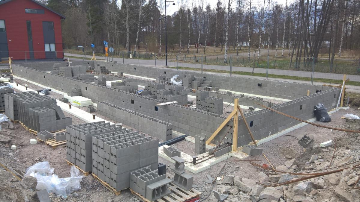 Rakennustyömaa, jossa harmaista betoniharkoista muurattu rakennuksen perustus. Etualalla on useita pinoja harkkoja puulavoilla. Taustalla vasemmalla on punainen talo ja oikealla tie, aita, katuvalaisin sekä paljaita puita. Kuva on otettu kirkkaassa päivänvalossa.
