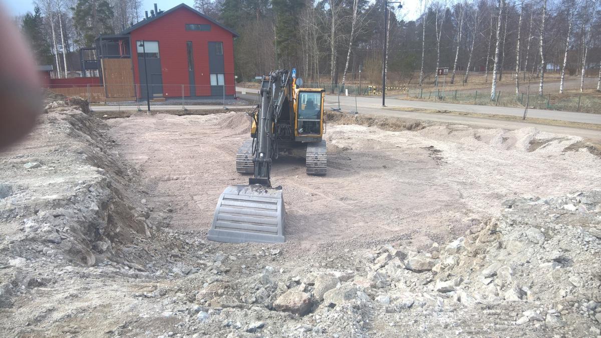 Keltainen kaivinkone seisoo rakennustyömaan keskellä tasaisella, vaalealla sora-alueella. Etualalla on louhittua kiveä ja maata. Taustalla vasemmalla näkyy moderni punainen talo tummanharmailla yksityiskohdilla aidan takana. Oikealla taustalla on tie ja rivi paljaita koivuja. Kuva on otettu aurinkoisena päivänä.