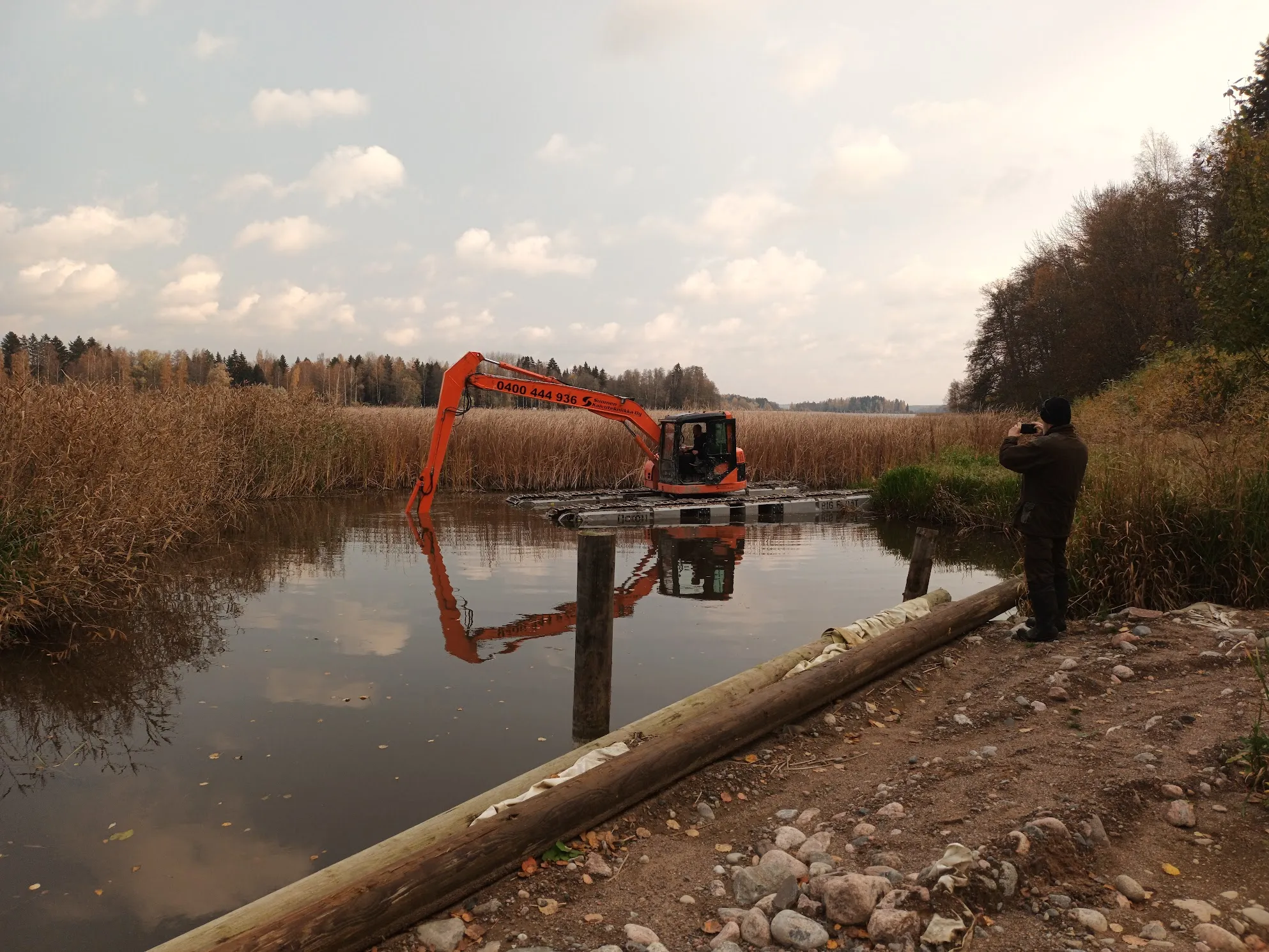 Oranssi kaivinkone kelluu vedessä kaislikon ympäröimänä pilvisenä päivänä. Oikealla rannalla tummiin pukeutunut henkilö valokuvaa kaivinkonetta puhelimellaan. Vesi heijastaa kaivinkoneen ja pilvisen taivaan. Rannalla on kiviä ja kaksi puupölkkyä.