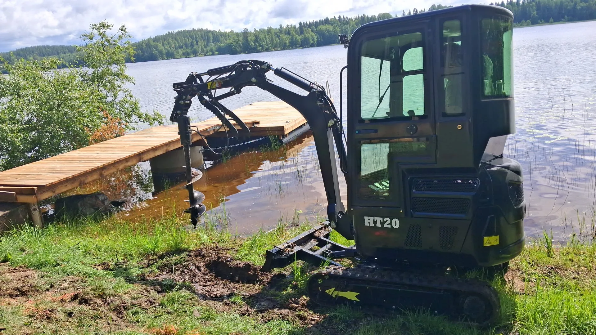 Musta HT20-minikaivuri maaporalla seisoo mutaisella, ruohoisella järvenrannalla. Kaivurin pora on veden äärellä, ja sen takana on puinen laituri tyynessä järvessä. Taustalla näkyy vihreä metsä ja pilvinen taivas.