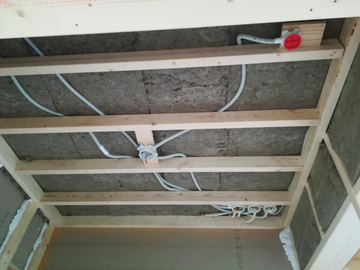 Kuvassa näkyy rakenteilla oleva katto, jossa on puinen koolaus ja sen välissä harmaata lämmöneristettä. Koolauksen päällä kulkee vaaleita sähköputkia ja useita jakorasioita. Yksi jakorasia on punakorkkinen ja toisaalla on kolme jakorasiaa rivissä. Alhaalla vasemmalla näkyy seinälevyä.