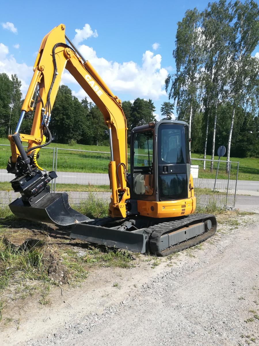 Keltainen ja harmaa Case-minikaivinkone seisoo sorapolulla vihreän nurmikon vieressä. Sen puomi on koholla ja kauha lepää maassa. Taustalla näkyy tie, puita ja sininen, pilvinen taivas kirkkaassa auringonvalossa.