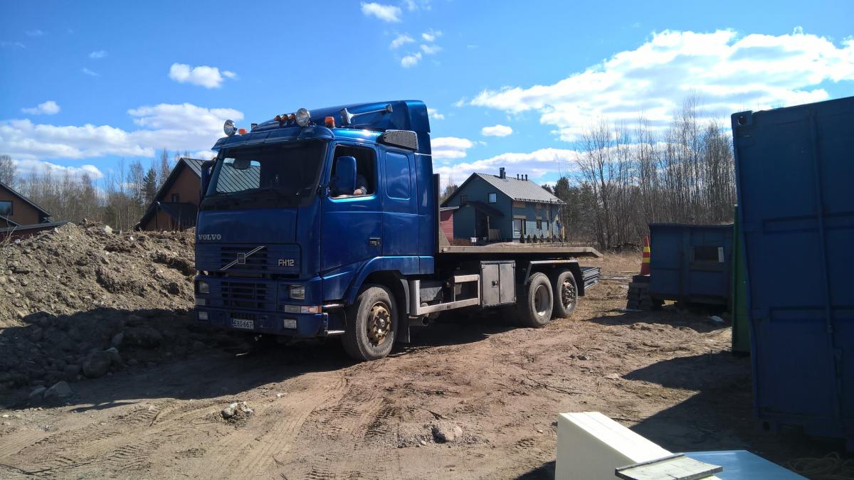 Sininen Volvo FH12 -kuorma-auto, jonka lavalla on pieni tummansininen talo, seisoo aurinkoisella hiekkaisella pihalla. Taustalla näkyy maakasa, puita ja muita rakennuksia sekä sininen merikontti.