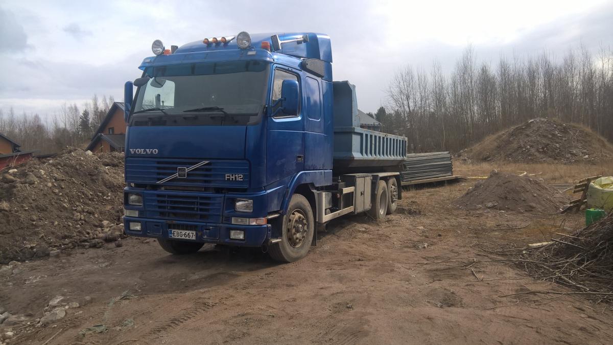 Sininen Volvo FH12 -kuorma-auto, jossa on kippilava, seisoo mutaisella maaperällä. Kuorma-auton ympärillä on maakasojen ja rakennustarvikkeiden pinoja. Taustalla näkyy paljaita puita ja pilvinen taivas. Vasemmalla etäällä on rakennuksia.