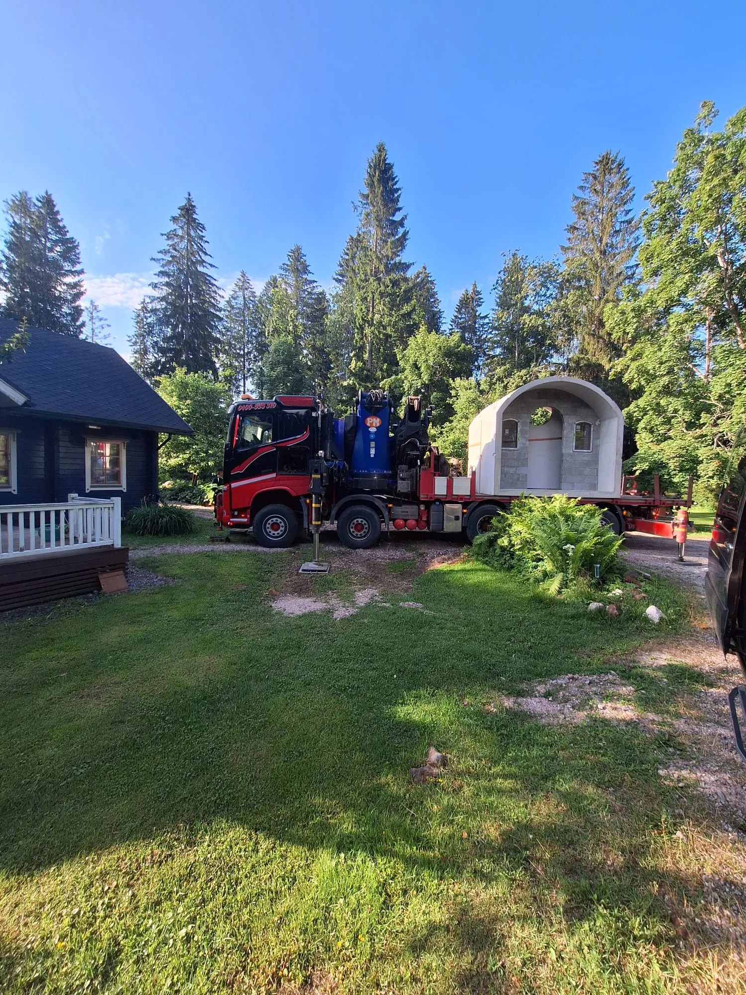 Aurinkoisena päivänä punamusta kuorma-auto, jossa on sininen nosturi, seisoo soratiellä tumman puutalon ja tiheän metsän edessä. Kuorma-auton lavalla on kaareva, esivalmistettu betonirakenne, jossa on kaksi ikkunaa ja pyöreä oviaukko. Etualalla on vihreää nurmikkoa ja saniaisia.