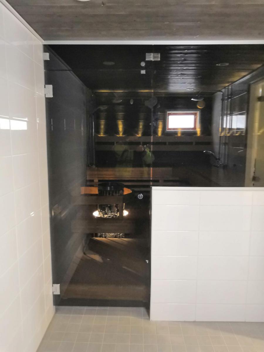 Kylpyhuone, jossa on lasiseinäinen sauna. Vasemmalla on valkoinen laattaseinä ja lasiovi. Oikealla on valkoinen laattapuoliseinä. Lattialla on vaaleanharmaat laatat ja katossa betonia sekä puupaneeleita. Saunan sisällä on tummat puupinnat, penkit ja useita ylöspäin suuntautuvia lämpimiä valaisimia. Pieni ikkuna päästää valoa sisään. Lasissa näkyy heijastus vihreäpaitaisesta henkilöstä ja suihkutilasta.