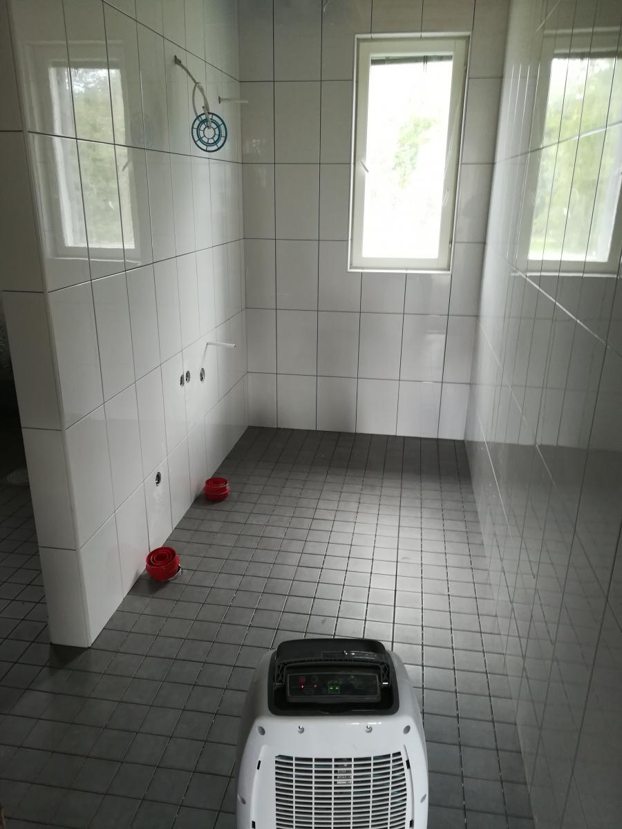 Huone, jossa on valkoiset seinälaatat ja tummanharmaat lattialaatat. Kaksi ikkunaa päästää sisään kirkasta luonnonvaloa. Seinillä näkyy putkia ja sininen pyöreä kiinnike. Lattialla on kaksi punaista putkenkorkkia ja etualalla valkoinen ilmankuivain, jonka näytössä palaa punaisia ja vihreitä valoja.