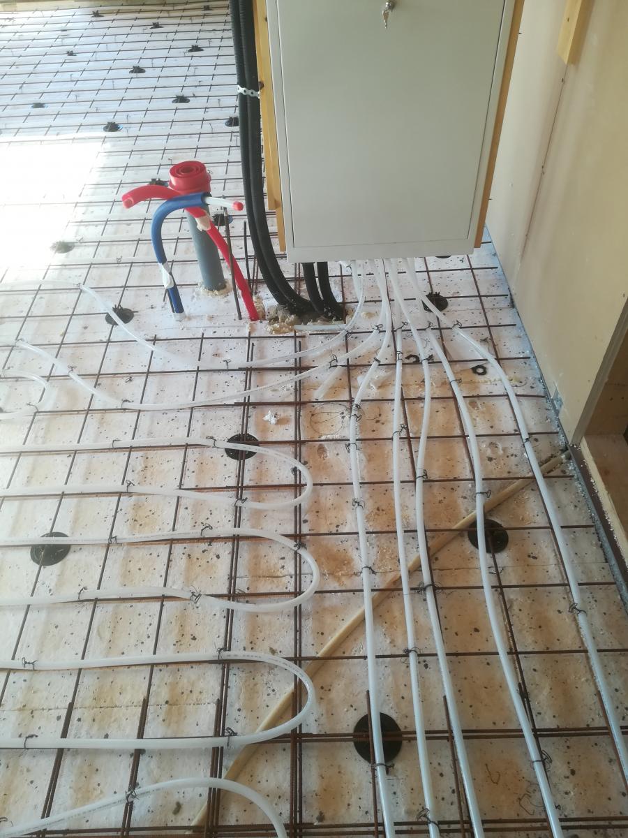 Lattialämmitysputkien asennus rakennustyömaalla. Valkoiset putket on kiinnitetty metalliverkkoon vaalean eristeen päällä. Oikealla ylhäällä on valkoinen kaappi, josta laskeutuu useita putkia ja kaapeleita lattiaan. Kaapin lähellä on myös punaisia, sinisiä ja mustia putkia sekä harmaa viemäriputki. Lattia on valmis betonivalua varten.