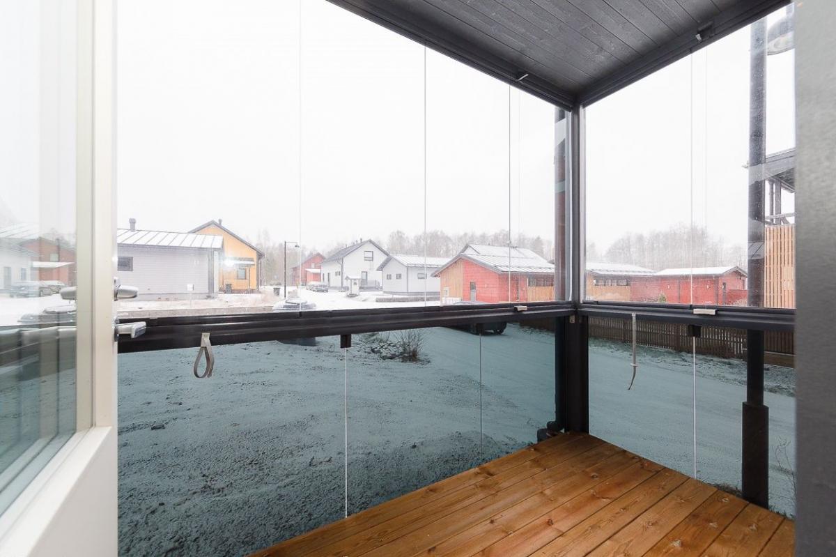 Näkymä lasitetulta parvekkeelta talvisena päivänä. Parvekkeella on vaaleanruskea puulattia ja tummat lasikehykset. Ulkona näkyy lumipeitteinen piha tai katu ja useita erivärisiä omakotitaloja, joiden katot ovat lumen peitossa. Taustalla on lumisia puita. Valo on tasainen ja pilvinen.
