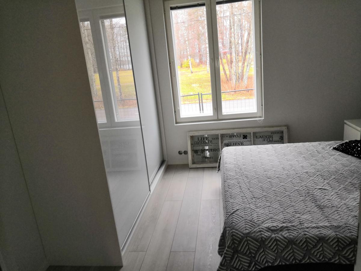 Makuuhuone, jossa on vaalea puulattia ja vaaleat seinät. Vasemmalla on suuri vaatekaappi peiliovilla, jotka heijastavat huonetta ja ikkunaa. Oikealla on sänky harmaalla, kuvioidulla päiväpeitolla ja tummalla tyynyllä. Takana on suuri ikkuna, josta näkyy ulos puita ja nurmikkoa pilvisenä päivänä. Ikkunan alla on valkoinen kehystetty taulu, jossa on tekstejä.