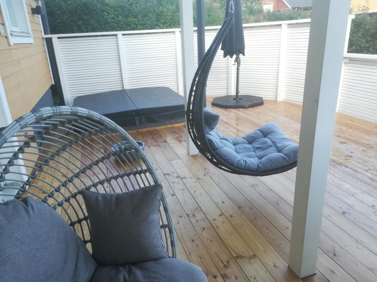 Märkä puuterassi, jossa vasemmalla harmaa pajutuoli harmailla tyynyillä ja oikealla musta riipputuoli vaaleansinisellä tyynyllä. Taustalla on musta poreallas kannella, suljettu aurinkovarjo ja valkoinen säleaita. Vasemmalla näkyy keltainen talon seinä ja taustalla vihreää kasvillisuutta.