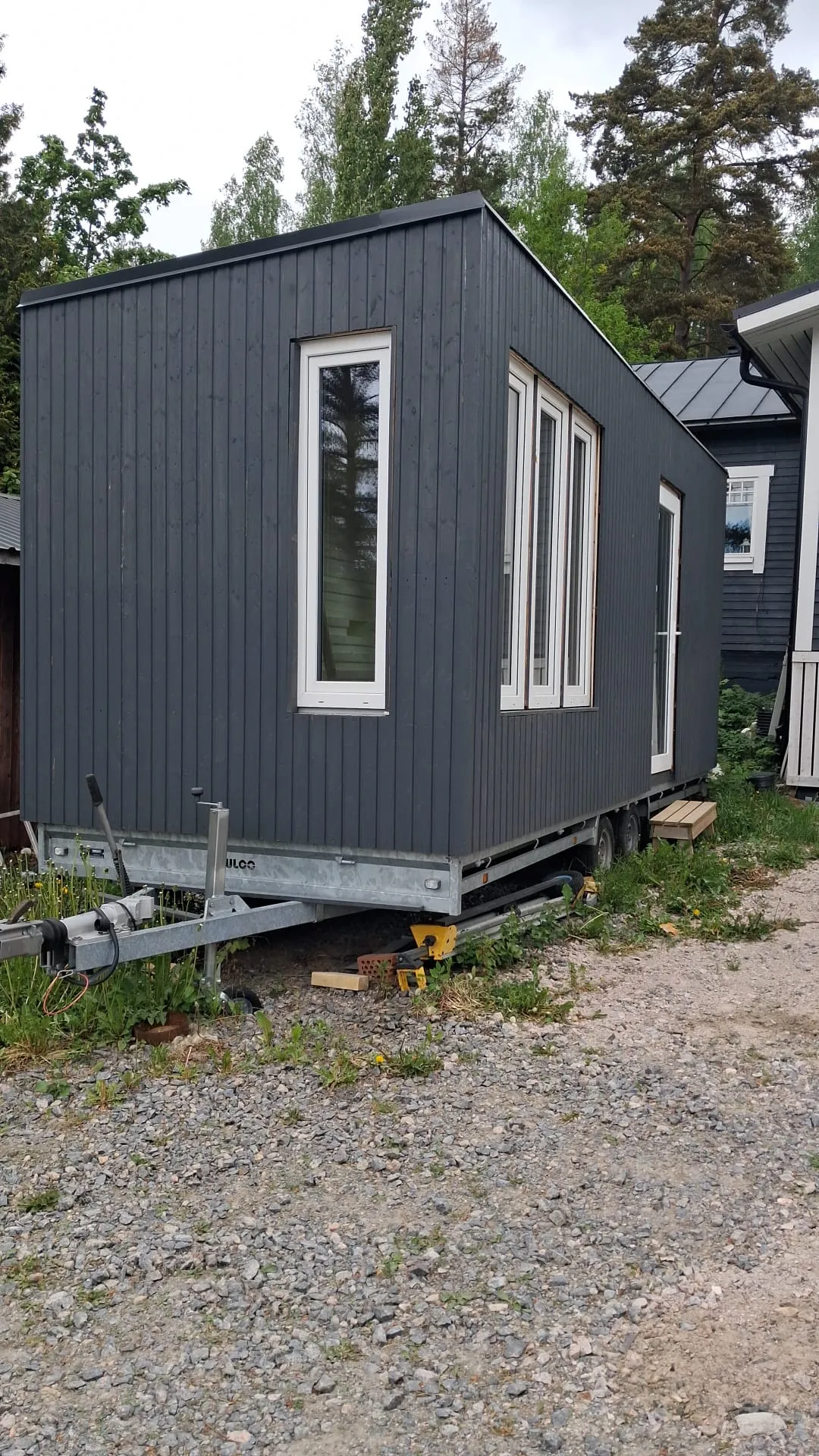 Tummanharmaa, pystylaudoitettu minitalo pyörillä seisoo soraisella pihalla. Talossa on valkoiset ikkunat ja valkoinen ovi. Perävaunun alusta ja pyörät ovat näkyvissä. Maassa on soraa, rikkaruohoja ja keltaisia tukia. Taustalla on vihreitä puita ja pilvinen taivas, sekä osittain toinen tumma rakennus.