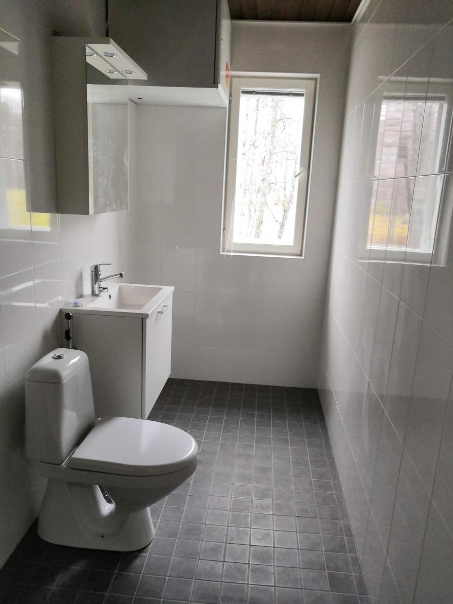 Valoisa, moderni wc-tila, jossa on valkoiset kaakeloidut seinät ja tummanharmaat lattialaatat. Vasemmalla on valkoinen wc-istuin ja sen vieressä valkoinen pesuallaskaappi. Pesualtaan yläpuolella on valkoinen peilikaappi, jossa on neljä valaisinta. Oikealla seinällä on kapea ikkuna, josta näkyy ulos puita, ja ikkunan heijastus näkyy vastakkaisella seinällä.