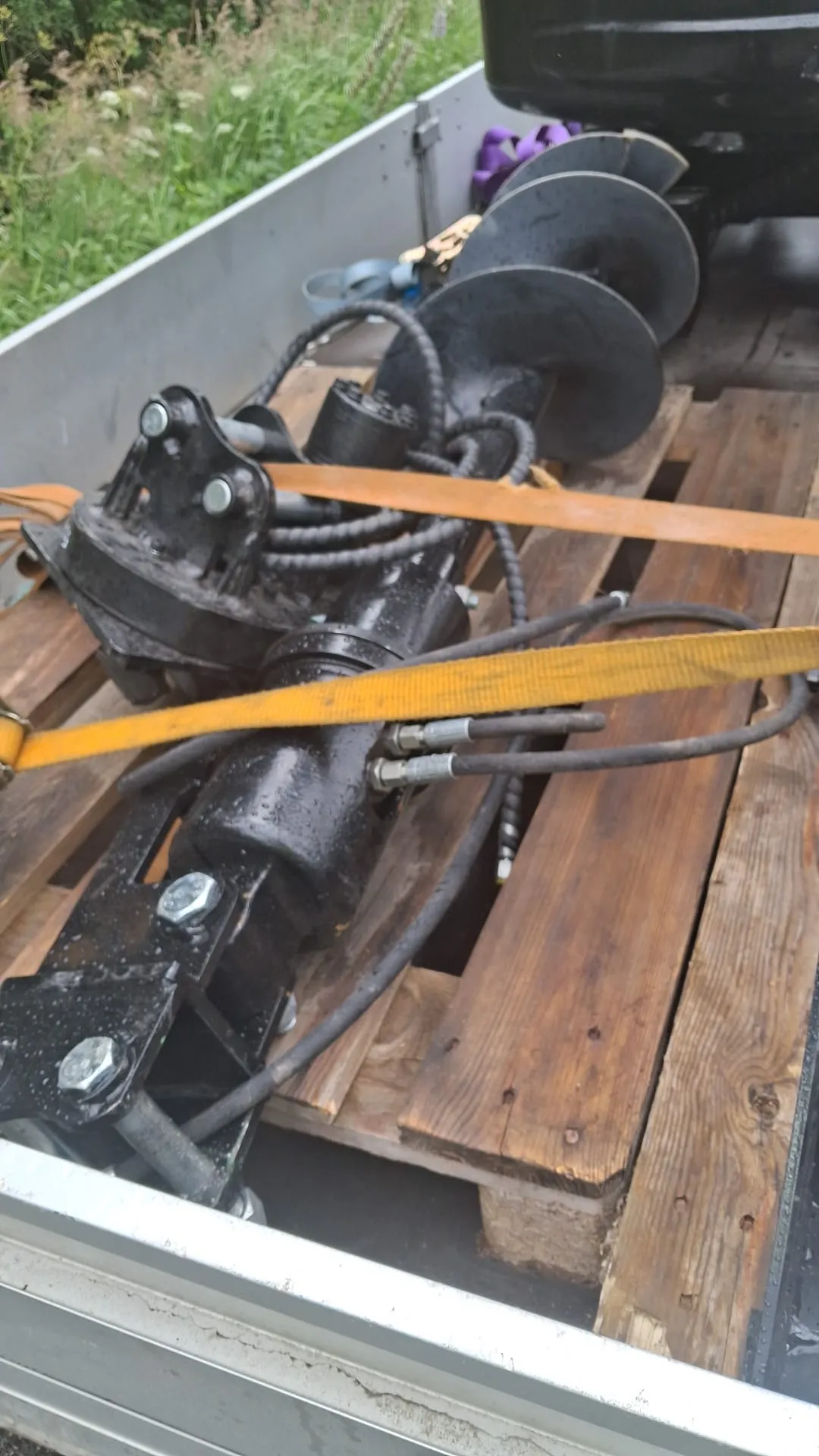 Musta, hydraulinen maapora tai ruuvipora, jossa on suuri kierreosa ja letkuja, makaa puisilla kuormalavoilla peräkärryn lavalla. Keltaiset kuormaliinat pitävät poraa paikoillaan. Laitteessa on vesipisaroita. Taustalla näkyy vihreää kasvillisuutta.