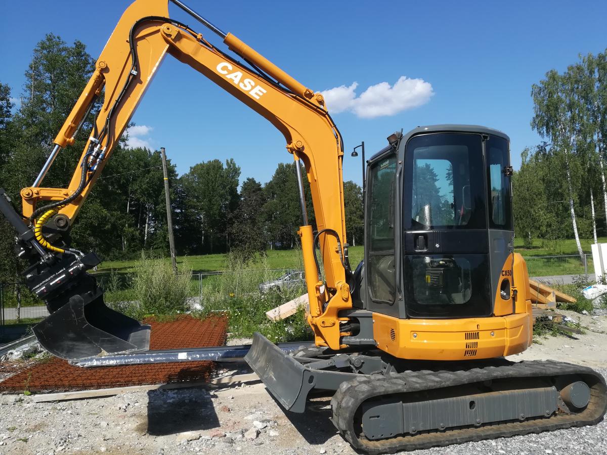 Oranssi ja harmaa Case CX50 -minikaivuri seisoo soraisella maalla aurinkoisena päivänä. Kaivurin puomi on nostettu ylös ja siinä on kiinni musta kauha. Taustalla näkyy puita ja sininen taivas valkoisine pilvineen.