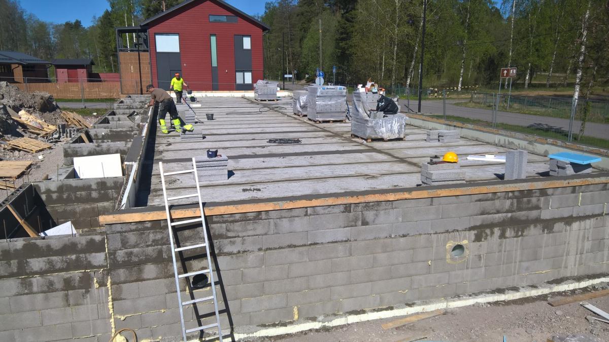Aurinkoisena päivänä otettu kuva rakennustyömaasta. Etualalla on harmaista betoniharkoista muurattu talon perustus ja sen päälle asennettu ontelolaattalattia. Kaksi rakennustyöntekijää työskentelee perustuksen parissa. Toinen on ruskeassa paidassa ja keltaisissa huomioväreissä housuissa, toinen keltaisessa paidassa. Lattialla on pinottuna rakennusmateriaaleja, keltainen kypärä ja sininen levy. Taustalla näkyy punainen, moderni talo ja vihreitä puita.