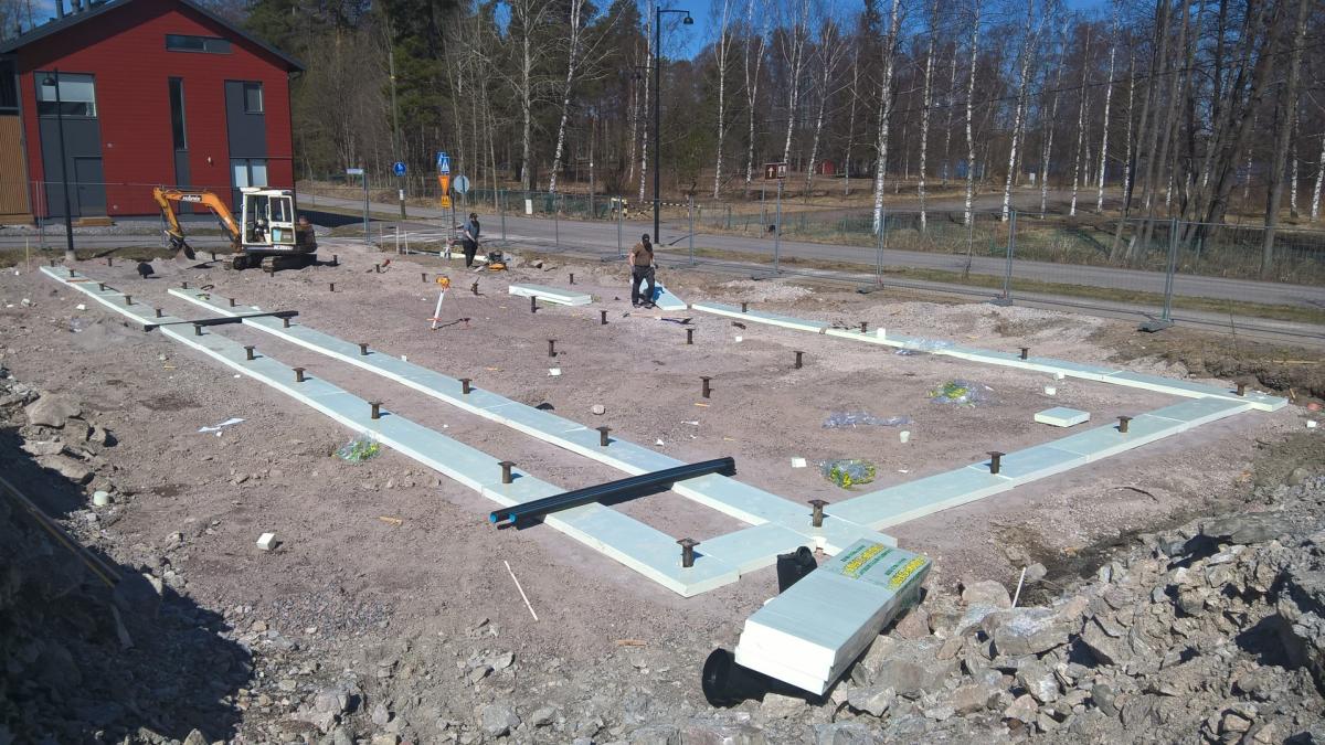 Aurinkoisella rakennustyömaalla valkoiset eristelevyt on aseteltu maahan muodostamaan rakennuksen perustuksen ääriviivat. Levyjen läpi nousee useita mustia T-muotoisia ankkureita. Vasemmalla on keltainen pienkaivuri ja kaksi työntekijää. Taustalla näkyy punainen rakennus, tie ja paljaita puita.