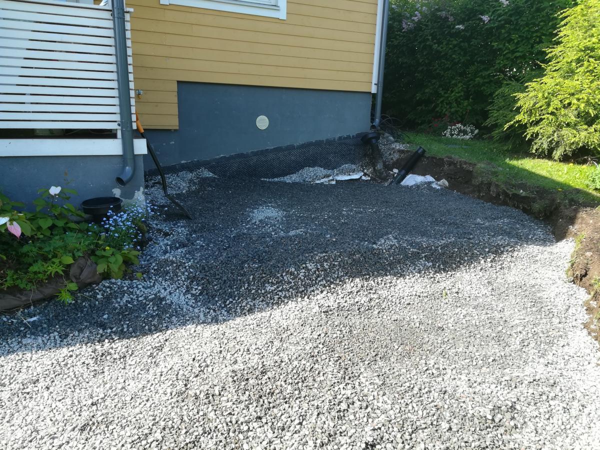 Kuva esittää talon pihaa, jossa on sepelöity alue ja maanrakennustöitä. Talon keltainen seinä ja tummanharmaa sokkeli näkyvät. Vasemmalla on valkoinen säleaita ja harmaa syöksytorvi. Etualalla on vaaleanharmaata sepeliä, ja talon vieressä tummempaa sepeliä, jonka alla on mustaa maisemointikangasta. Oikealla on kaivettu oja, ja taustalla on vihreää nurmikkoa ja pensaita. Talon vieressä on musta lapio oranssilla varrella ja musta ämpäri kasvien keskellä. Aurinko paistaa kirkkaasti, luoden varjoja sepelipinnalle.