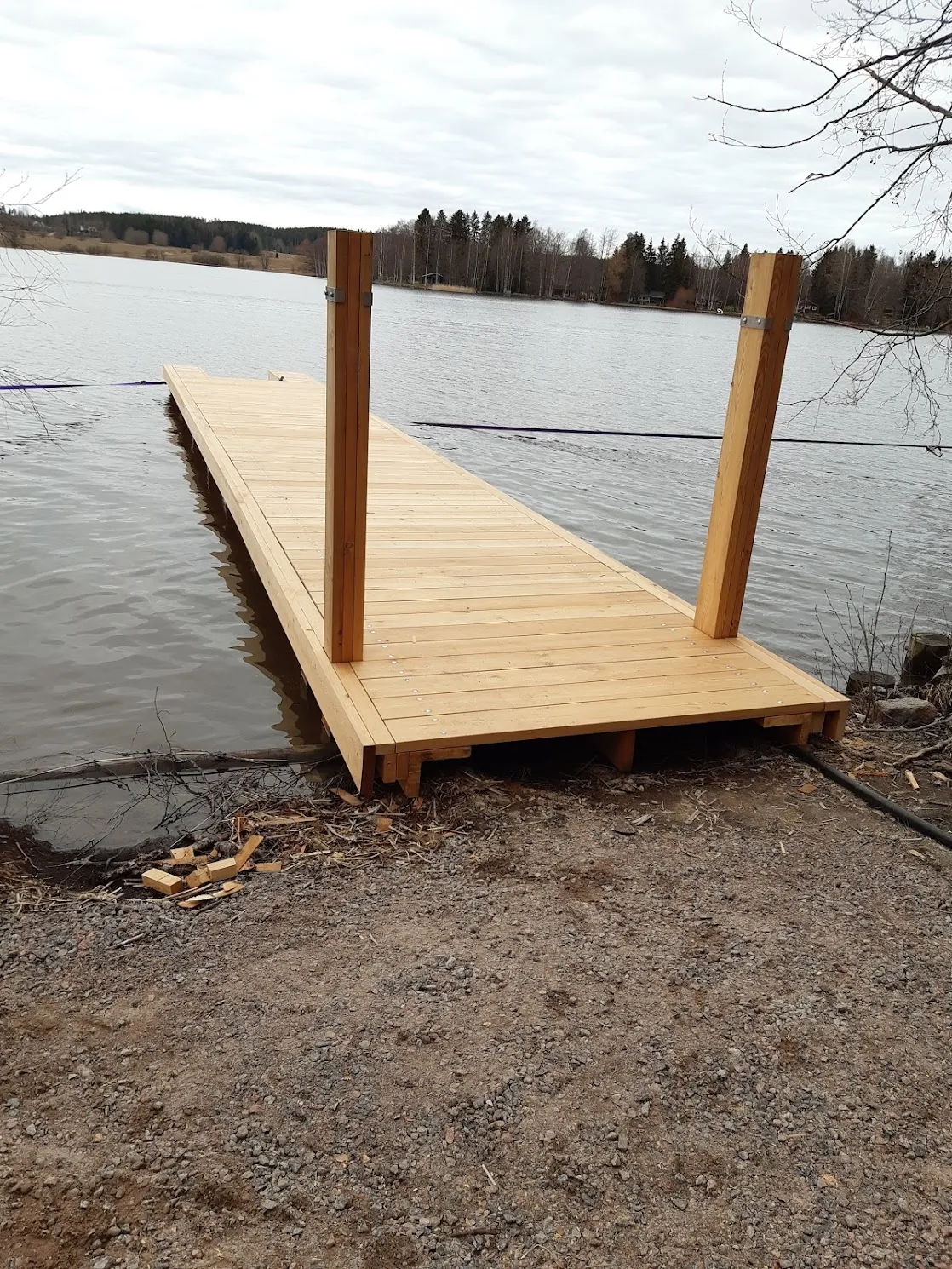 Uusi vaaleapuinen laituri ulottuu järveen pilvisenä päivänä. Laiturin maapuolella on kaksi pystysuoraa puutolppaa. Ranta on sorainen, ja veden rajassa on puujätettä. Taustalla näkyy metsäinen vastaranta.