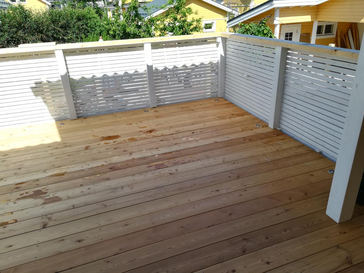 Vaaleanruskea puinen terassi, jossa on märkiä läiskiä ja aurinkoisia kohtia. Terassia ympäröi valkoinen säleaita, jonka varjot näkyvät lattialla. Taustalla on keltaisia taloja ja vihreää kasvillisuutta.