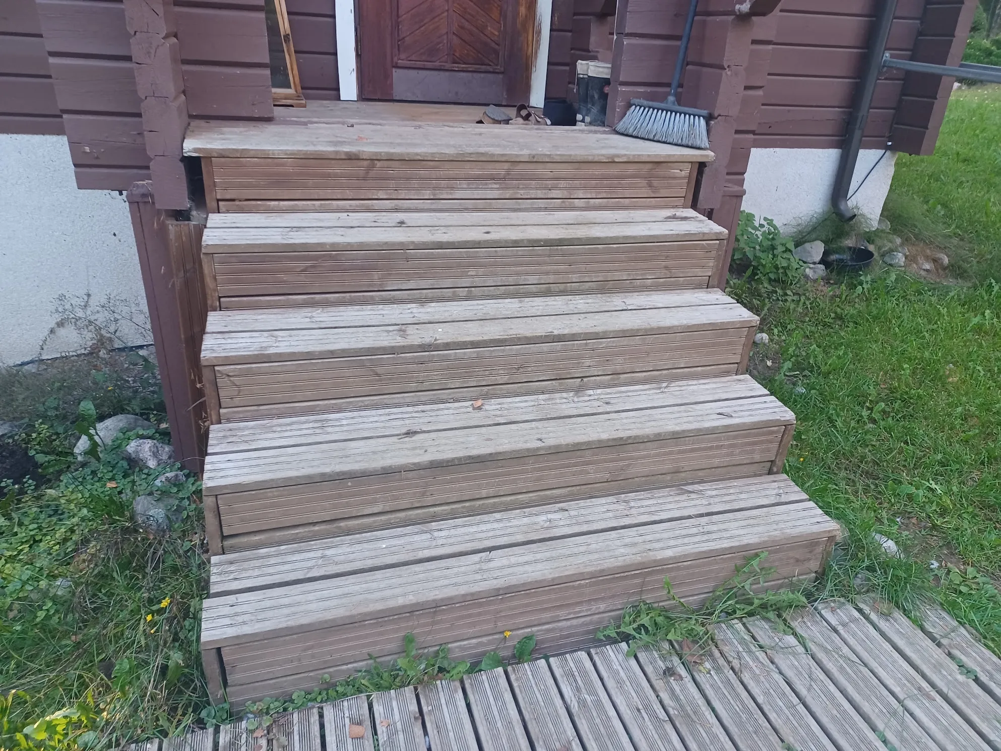 Kuvassa on viisi puiset askelmaa, jotka johtavat ruskean puutalon sisäänkäynnille. Portaiden puu on vaaleanruskeaa ja säänkestävää. Talon ruskea seinä ja valkoinen perustus näkyvät. Oikealla portaiden vieressä on musta harja ja vasemmalla vihreää kasvillisuutta. Portaiden edessä on puinen terassi ja ympärillä vihreää nurmikkoa.