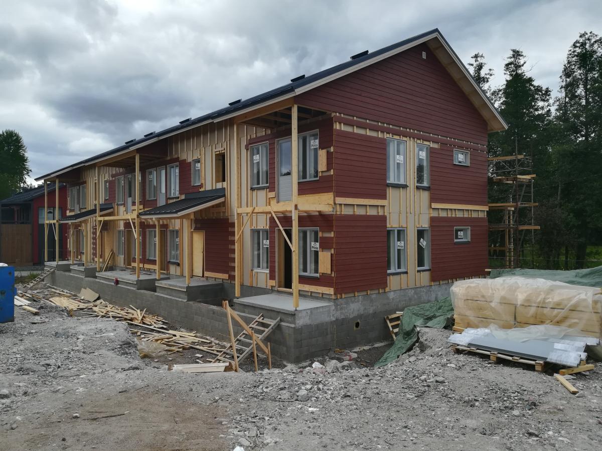 Kuva esittää rakenteilla olevaa rivitaloa pilvisenä päivänä. Rakennuksen julkisivussa on tummanpunaista puuverhousta ja osittain näkyvää puurunkoa. Ikkunat on asennettu. Etualalla on rakennustyömaa, jossa on soraa, puunpalasia ja rakennustarvikkeita, kuten muoviin käärittyä puutavaraa ja lasilevyjä lavoilla. Taustalla näkyy puita ja rakennustelineitä.