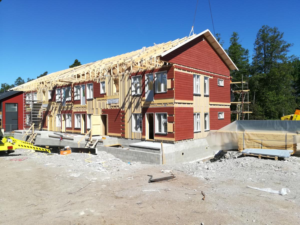 Aurinkoisena päivänä otettu kuva rakenteilla olevasta, punaisella ulkoverhouksella ja valkoisilla ikkunoilla varustetusta rakennuksesta. Rakennuksen yläosa ja katto ovat vielä puurunkoisia. Taustalla näkyy vihreitä puita ja kirkas sininen taivas. Etualalla on rakennustarvikkeita ja soraa.