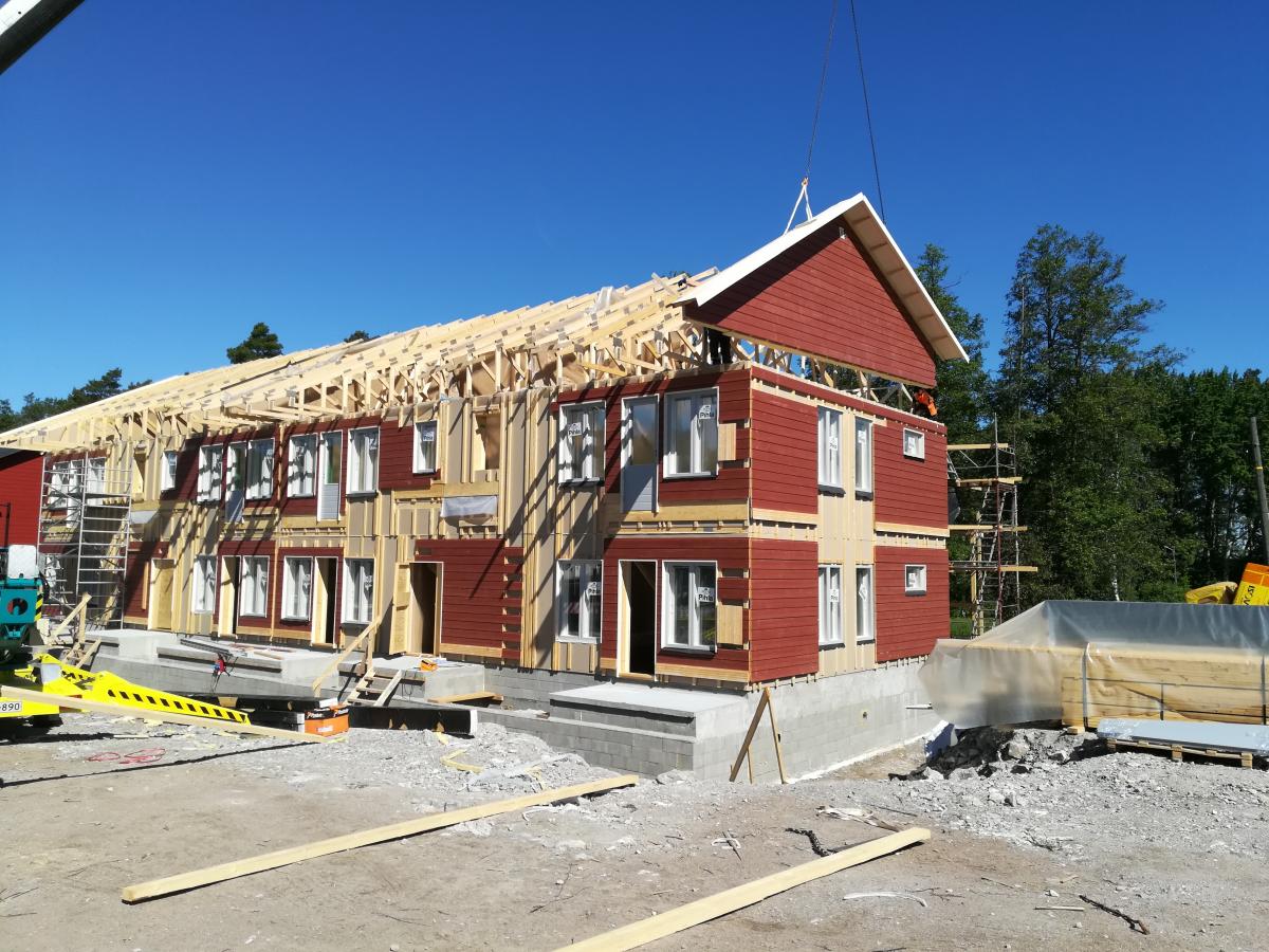 Aurinkoisena päivänä rakenteilla oleva kaksikerroksinen rakennus. Sen alaosassa on punainen ulkoverhous ja valkoiset ikkunat. Yläosassa näkyy puinen kattorunko, ja nosturi nostaa punaista päätykolmiota paikoilleen. Taustalla on kirkas sininen taivas ja vihreitä puita. Maassa on rakennustyömaan materiaaleja.