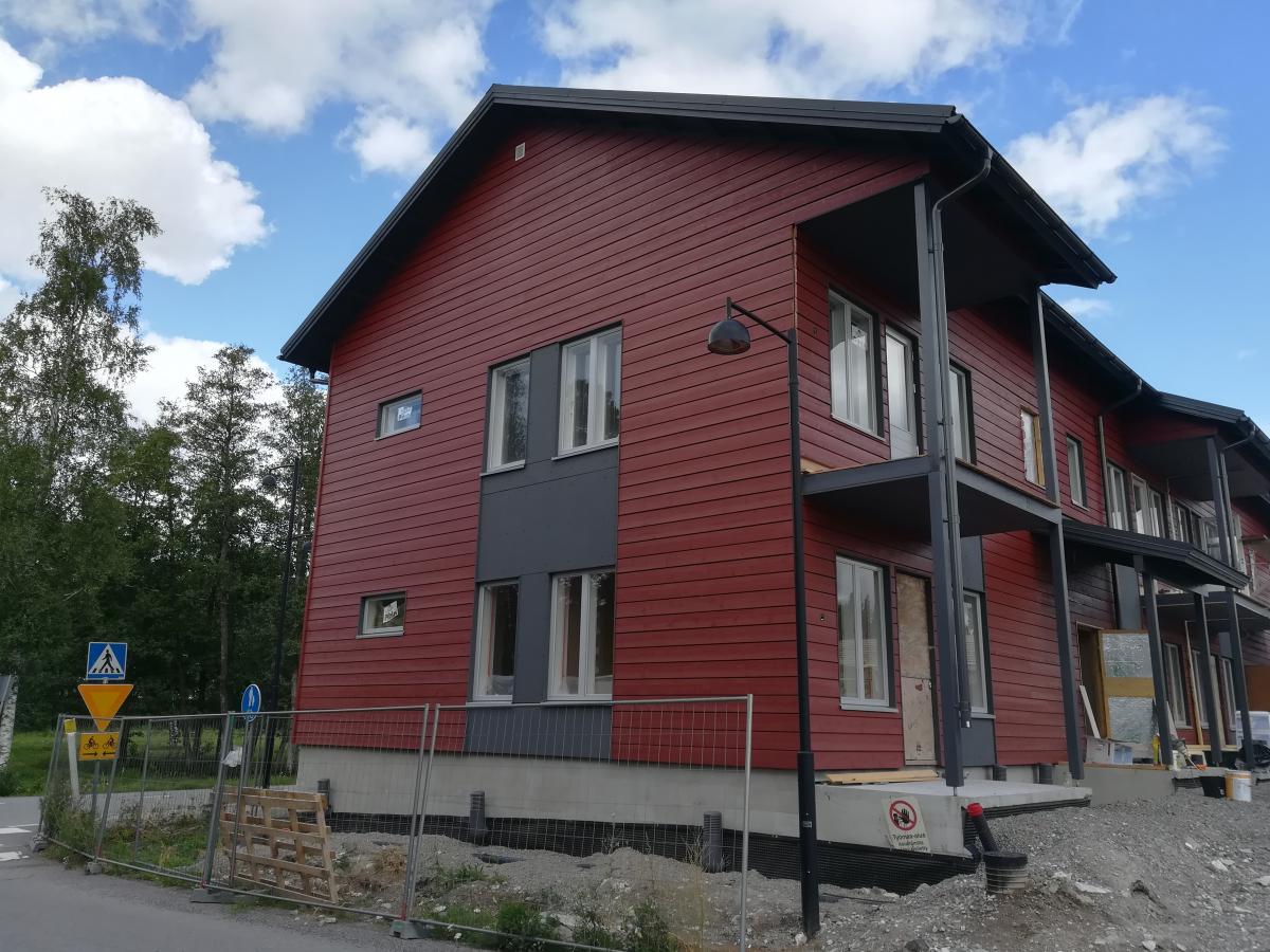 Punainen, kaksikerroksinen puurakennus on rakenteilla aurinkoisena päivänä. Rakennuksessa on tummanharmaat parvekkeet ja valkoiset ikkunanpuitteet. Edustalla on rakennustyömaa-aita, puulava ja soraa. Vasemmalla näkyy katulamppu ja liikennemerkki, jossa on suojatie- ja pyöräilysymbolit. Taustalla on vihreitä puita ja sininen taivas valkoisilla pilvillä. Rakennuksen sokkelissa on punainen kieltokyltti tekstillä "Työmaa-alue Pääsy kielletty".