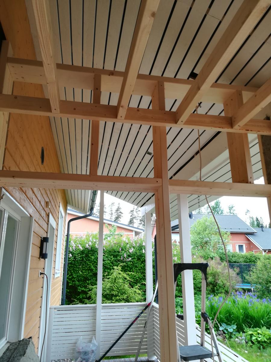 Kuva esittää osittain valmista puista kuistia. Katossa on vaaleita puulautoja tummilla raidoilla ja näkyvissä on vaalea puinen kattorakenne. Vasemmalla on talon keltainen puuseinä valkoisella ovella ja mustalla ulkovalolla. Oikealla ja edessä on vaaleita puupylväitä ja -palkkeja. Alhaalla on valkoinen sälekaide. Kaiteen takana näkyy vehreä puutarha pensaineen, puineen ja sinisine kukkineen. Taustalla on taloja tummilla katoilla. Kuvassa on myös tikkaat ja roikkuva johto. Valo on luonnollinen ja hajanainen.