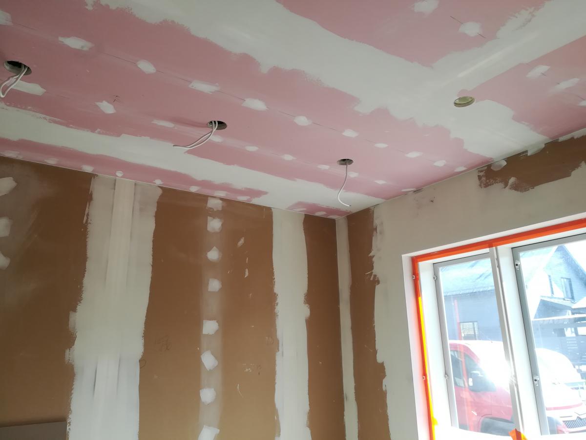 Rakenteilla oleva huone, jossa katto on levytetty ja tasoitettu osittain vaaleanpunaisella tasoitteella valkoisten levyjen päällä. Katossa on useita reikiä sähköjohtoineen valaisimia varten. Seinät ovat ruskeaa kipsilevyä, jossa on valkoisia tasoitevetoja saumojen ja ruuvien päällä. Oikealla on suuri ikkuna oransseilla karmeilla, josta näkyy ulos punainen pakettiauto ja rakennuksia.