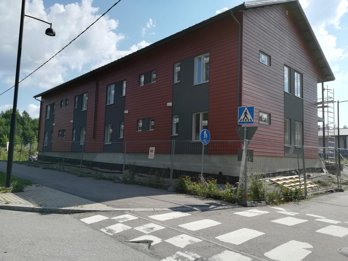 Kaksikerroksinen punaruskea ja tummanharmaa rakennus aurinkoisena päivänä. Rakennuksen ympärillä on metallinen rakennusaita ja oikealla puolella rakennustelineitä. Kuvassa näkyy kadun risteys suojateineen ja vasemmalla katuvalopylväs. Aidalla on kaksi sinistä liikennemerkkiä: suojatie ja jalankulkutie. Taustalla on puita ja pilvinen taivas.