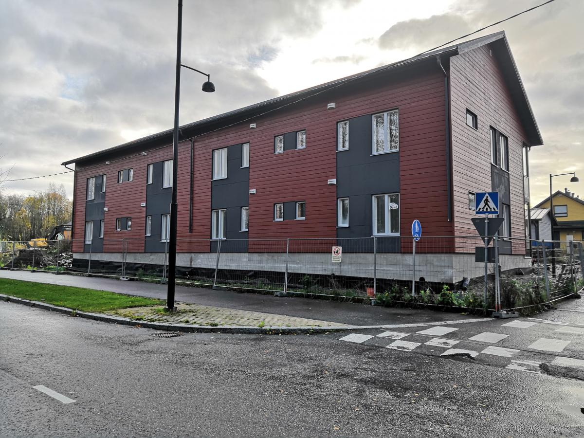 Kaksikerroksinen rakennus, jossa on punaruskea puuverhoilu ja tummanharmaat ikkunapaneelit, seisoo pilvisenä päivänä. Edustalla on asfalttitie suojatiellä, musta katuvalopylväs ja liikennemerkkejä. Rakennuksen ympärillä on metallinen työmaa-aita, ja taustalla näkyy puita sekä keltainen kaivuri.