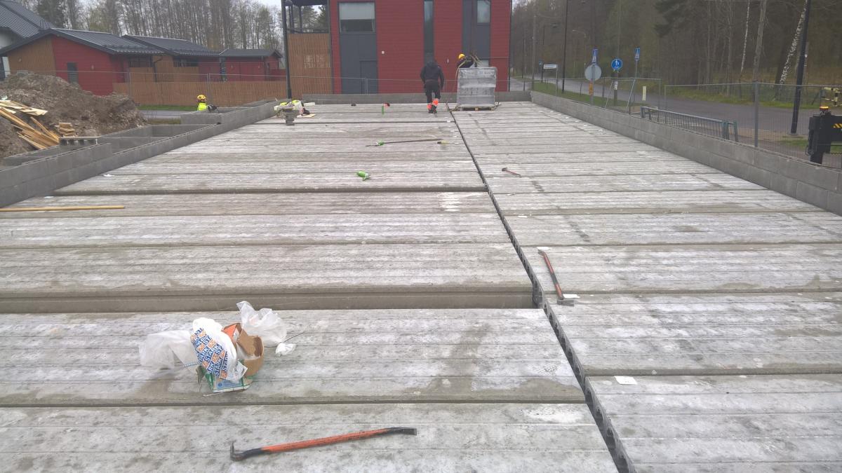 Rakennustyömaa pilvisenä päivänä. Etualalla on laaja alue harmaita ontelolaattoja, joissa on rakoja. Laatoilla näkyy roskia ja työkalut, kuten sorkkarauta. Keskellä useita rakennustyöntekijöitä, joista yksi kävelee punaista rakennusta kohti. Vasemmalla on maakasa ja punaisia rakennuksia. Oikealla näkyy tie, liikennemerkkejä ja metalliaita. Valo on tasainen.