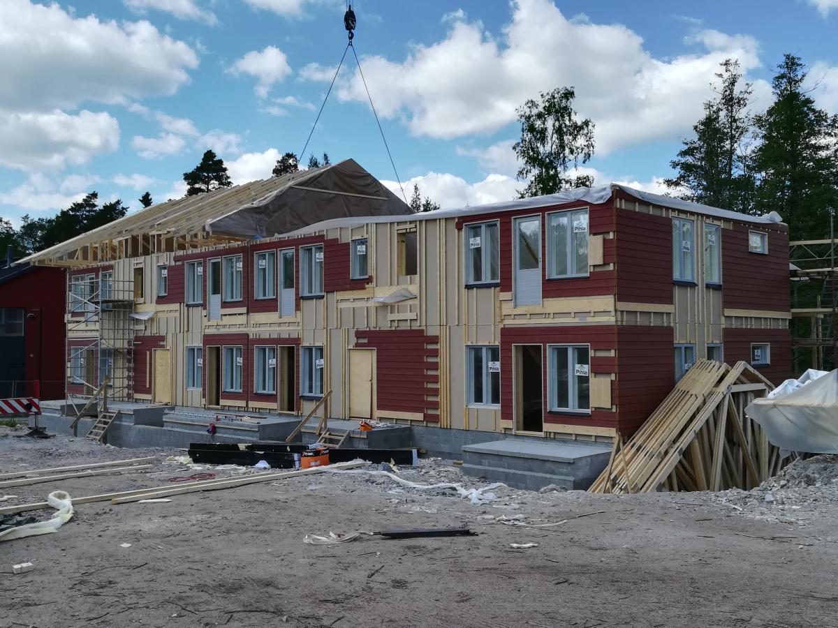 Rakennustyömaa aurinkoisena päivänä. Kaksikerroksinen rakennus on rakenteilla, sen seinissä on punaista puuverhousta ja paljasta puurunkoa. Ikkunat ovat asennettuina. Nosturi nostaa kattorakennetta rakennuksen vasempaan osaan. Taustalla on vihreitä puita ja sininen taivas valkoisine pilvineen.