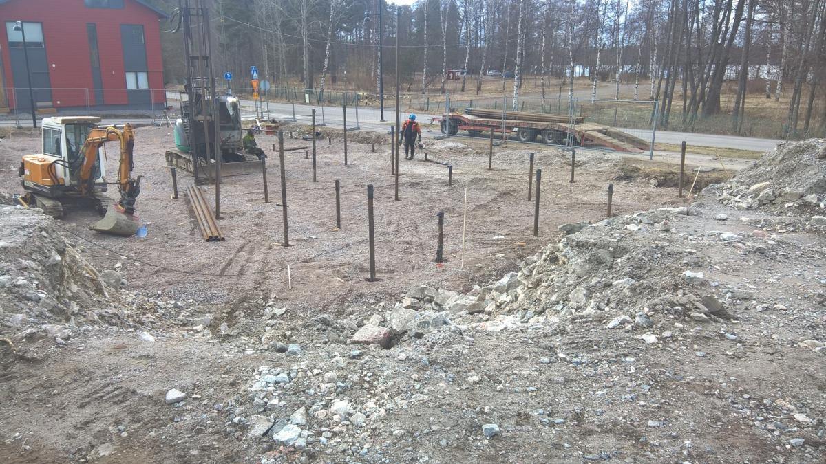 Rakennustyömaa, jossa maahan on asennettu useita tummia paaluja. Vasemmalla on keltainen kaivinkone ja oikealla paalutuskone, jonka päällä istuu työntekijä. Toinen työntekijä seisoo paalujen joukossa. Maassa on soraa, kiviä ja kaivettua maata. Taustalla näkyy punainen rakennus, tie ja lehdettömiä puita.