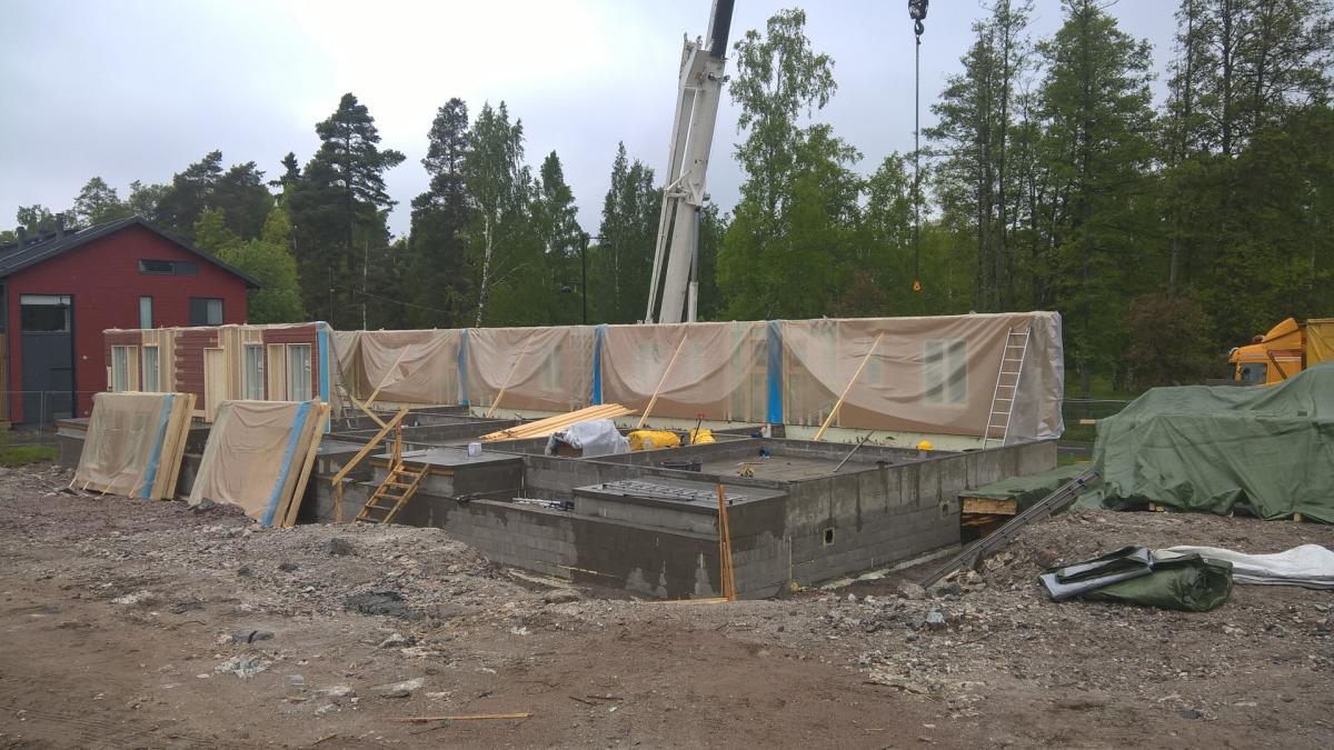 Rakennustyömaa, jossa on uuden talon betoninen perustus. Vasemmalla punainen talo ja keskellä pystyssä vaaleilla pressuilla suojattuja seinäelementtejä. Taustalla näkyy nosturin puomi ja tiheää metsää. Etualalla on kivistä ja mutaista maata. Oikealla on vihreillä pressuilla peitettyjä rakennustarvikkeita ja keltainen kuorma-auto. Kuva on otettu pilvisenä päivänä.