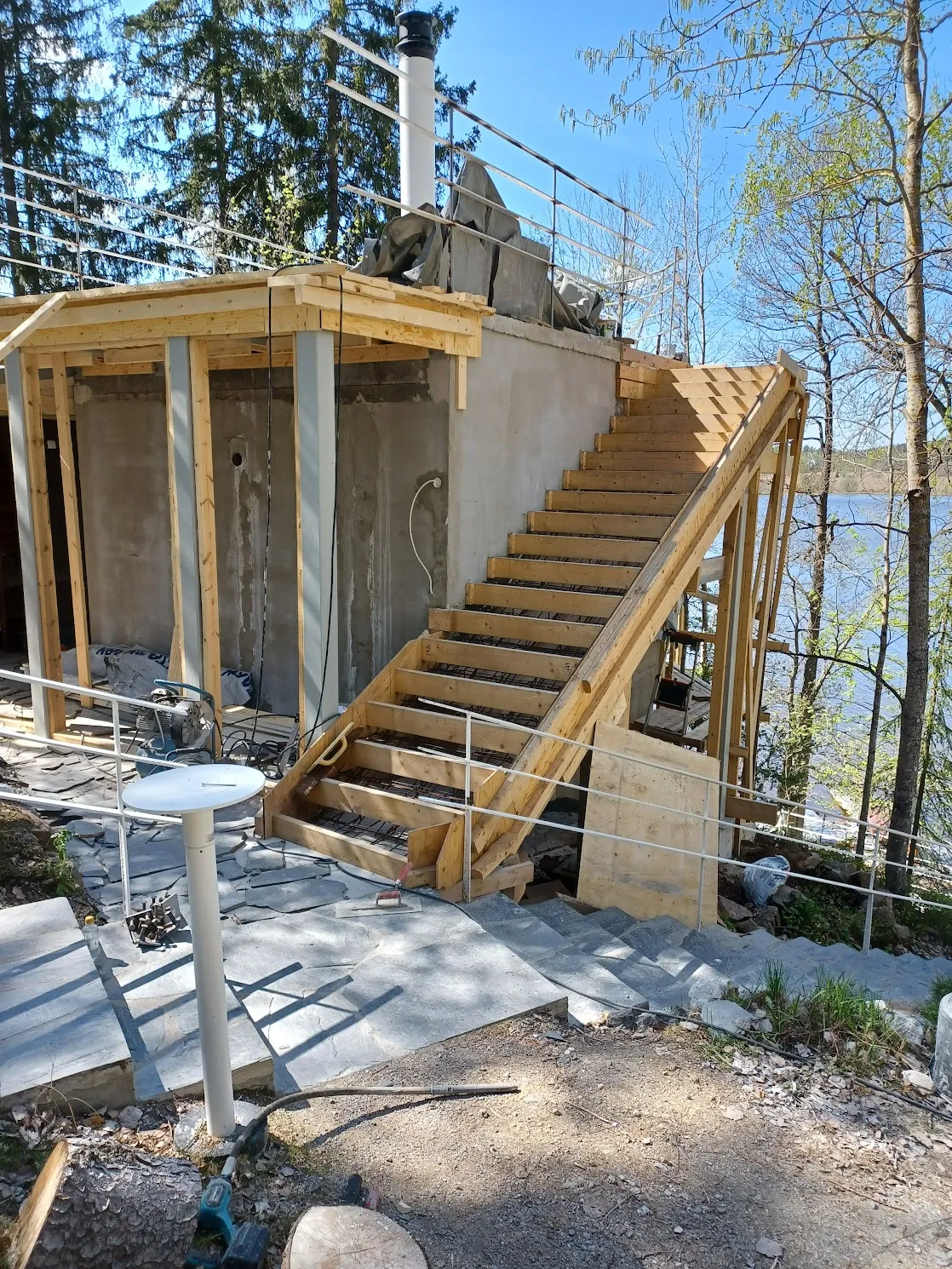 Kuva esittää keskeneräistä rakennusta järven rannalla aurinkoisena päivänä. Rakennuksessa on harmaat betoniseinät ja puurunkoiset portaat, joissa on metallikaiteet. Portaat johtavat ylös rakennuksen tasanteelle, jolla on valkoinen savupiippu tai ilmastointiputki. Taustalla näkyy sininen järvi ja vihreitä puita. Etualalla on harmaata kivetystä, ruskeaa maaperää ja valkoinen ulkopöytä.