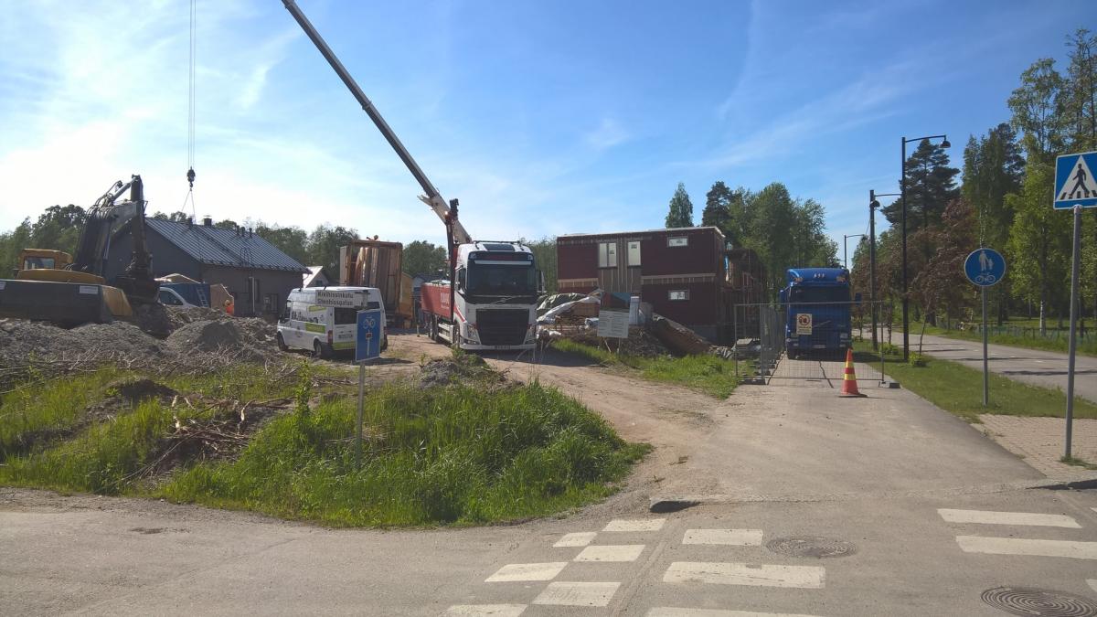 Aurinkoisena päivänä kuvattu rakennustyömaa. Vasemmalla kaivinkone ja maakasat, ja nosturi nostaa esinettä tummakattoisen rakennuksen ylle. Keskellä valkoinen ja punainen kuorma-auto nosturilla, sekä modulaarisia rakennusosia. Oikealla sininen kuorma-auto, rakennusaitoja, oranssi kartio ja jalkakäytävä suojatiellä. Taustalla puita ja kirkas sininen taivas.