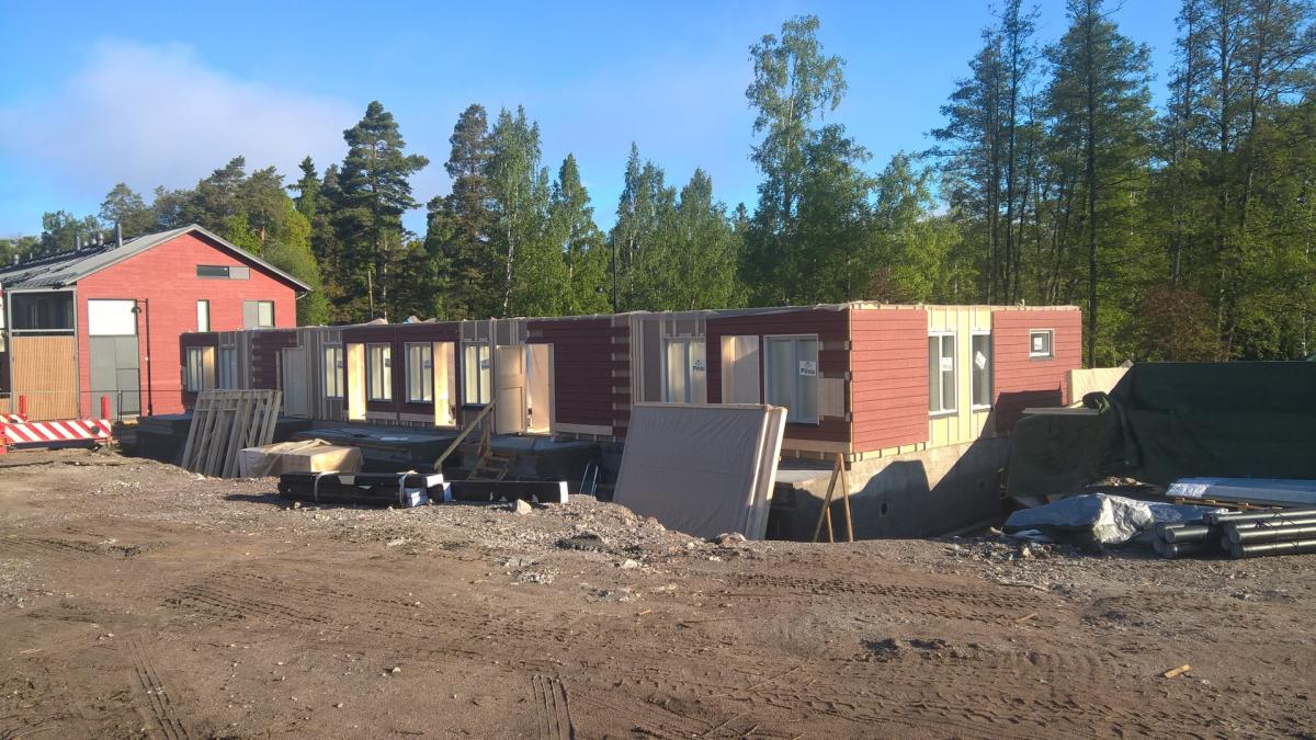 Aurinkoinen kuva rakennustyömaasta, jossa vasemmalla on punainen, valko-harmailla yksityiskohdilla varustettu talo ja oikealla rakenteilla oleva, punaisella ulkoverhouksella ja vaaleilla seinillä varustettu talo. Etualalla on hiekkainen maa, jossa näkyy renkaanjälkiä ja erilaisia rakennustarvikkeita, kuten puutavaraa, eristelevyjä ja putkia. Taustalla kohoaa tiheä vihreä metsä ja yläpuolella on kirkas sininen taivas muutamine valkoisine pilvineen.