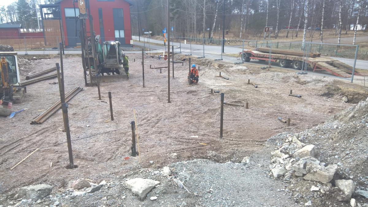 Rakennustyömaa pilvisenä päivänä. Keskellä kuvaa on porauskone ja useita maahan upotettuja metallipylväitä. Kaksi työntekijää työskentelee: toinen oranssissa takissa ja sinisessä kypärässä kyykistyneenä, toinen vihreässä takissa porauskoneen vieressä. Vasemmalla on kaivinkone ja maassa pino putkia. Taustalla näkyy punainen rakennus, tie, puita ja peräkärry täynnä putkia aidan takana. Maa on soraa ja likaa.