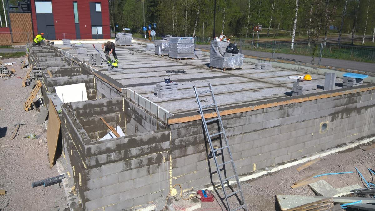 Aurinkoisena päivänä rakennustyömaalla kaksi työntekijää työskentelee suuren harmaan betoniharkkoperustuksen päällä. Perustuksen yläosa on betonilaatta, jolla on pinottuna rakennusmateriaaleja ja työkaluja. Oikealla nojaa tikkaat perustusta vasten. Taustalla näkyy punainen rakennus, puita ja tie.