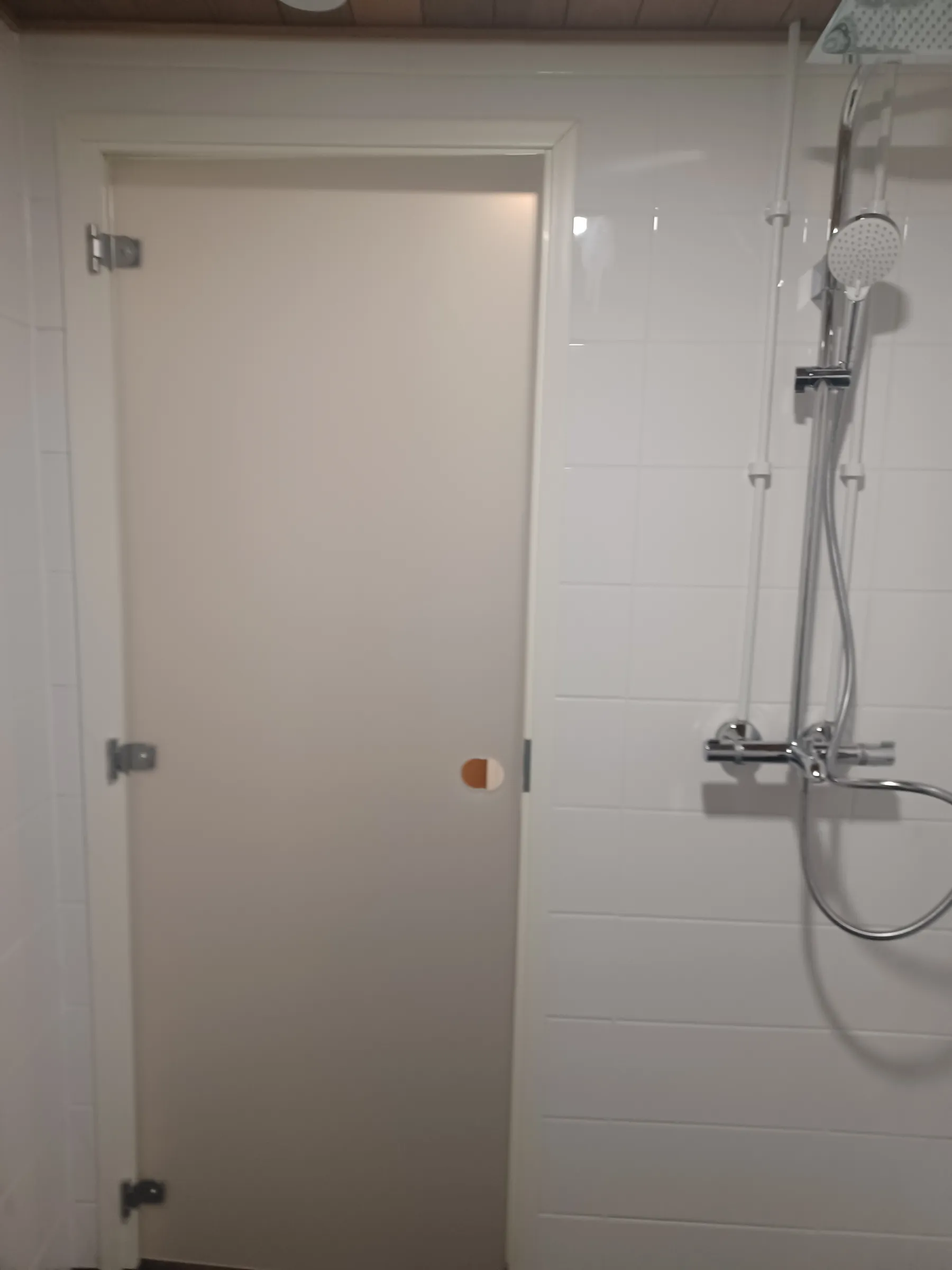 Kuva esittää kylpyhuoneen tai saunan sisätilaa. Vasemmalla on vaalea, himmennetty lasiovi, jossa on kolme hopeista saranaa ja pyöreä, kaksivärinen vedin. Oikealla on valkoisilla laatoilla päällystetty seinä, johon on asennettu krominen suihkujärjestelmä käsisuihkulla ja yläsuihkulla. Yläreunassa näkyy puupaneelikatto.